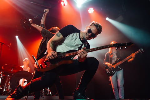 Guitarist is rocking out on stage, playing with intensity and passion under the glow of red stage lights, surrounded by his bandmates who are also giving their all to the performance