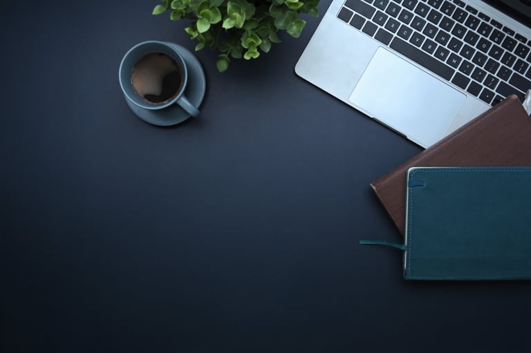 Top view of laptop, coffee cup, notebooks, and green plant on dark desk.